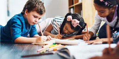 Trois enfants allongés sur le ventre par terre, faisant du coloriage avec des crayons de couleur.