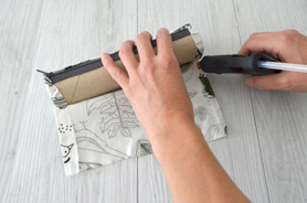 Mains en train de coller un tissu autour d’un tube de rouleau d’essuie-tout à l’aide d’un pistolet à colle.