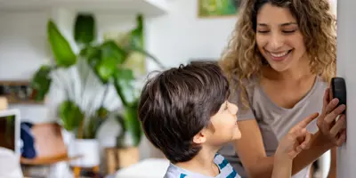 Une mère aux cheveux bouclés et un jeune garçon souriant changent un thermostat.